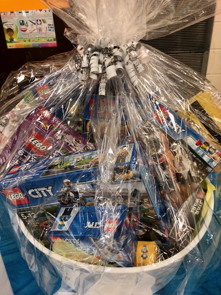 Photo of a dream basket from a past year; it's a container wrapped in clear cellophane and tied with a bow that is filled with Lego goodies