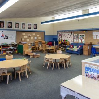 A large classroom with small tables and small chairs, a grey carpet and light-coloured walls covered with bulletin boards and a white board with school-related items posted on them.
