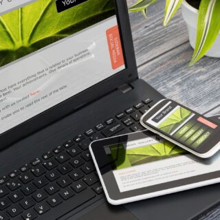 An open laptop with a white and green page displayed on the screen. A tablet and cellphone are piled on the laptop keyboard and there are bits of a potted plant visible in the top right corner.